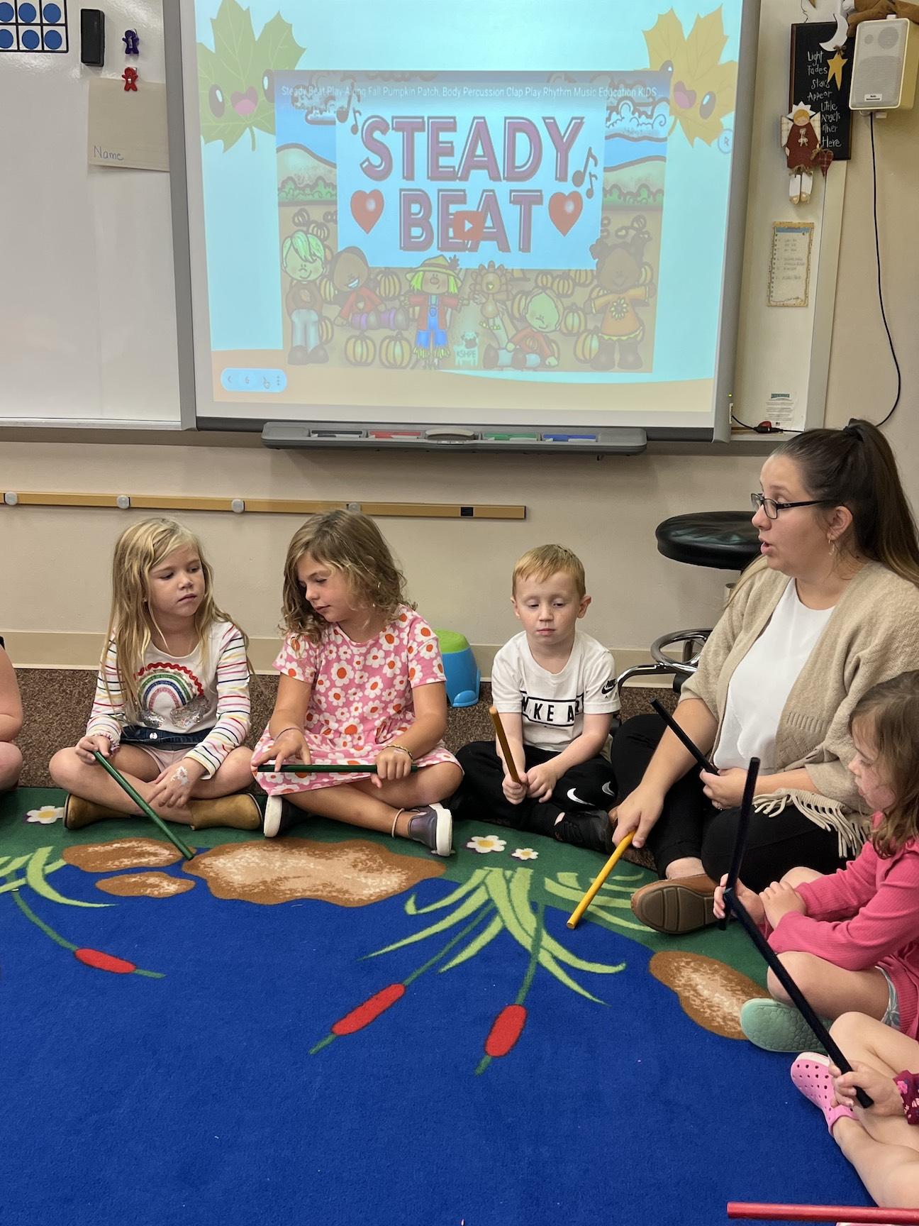 Mrs. Olive leads the music lesson by demonstrating a steady beat with the rhythm sticks